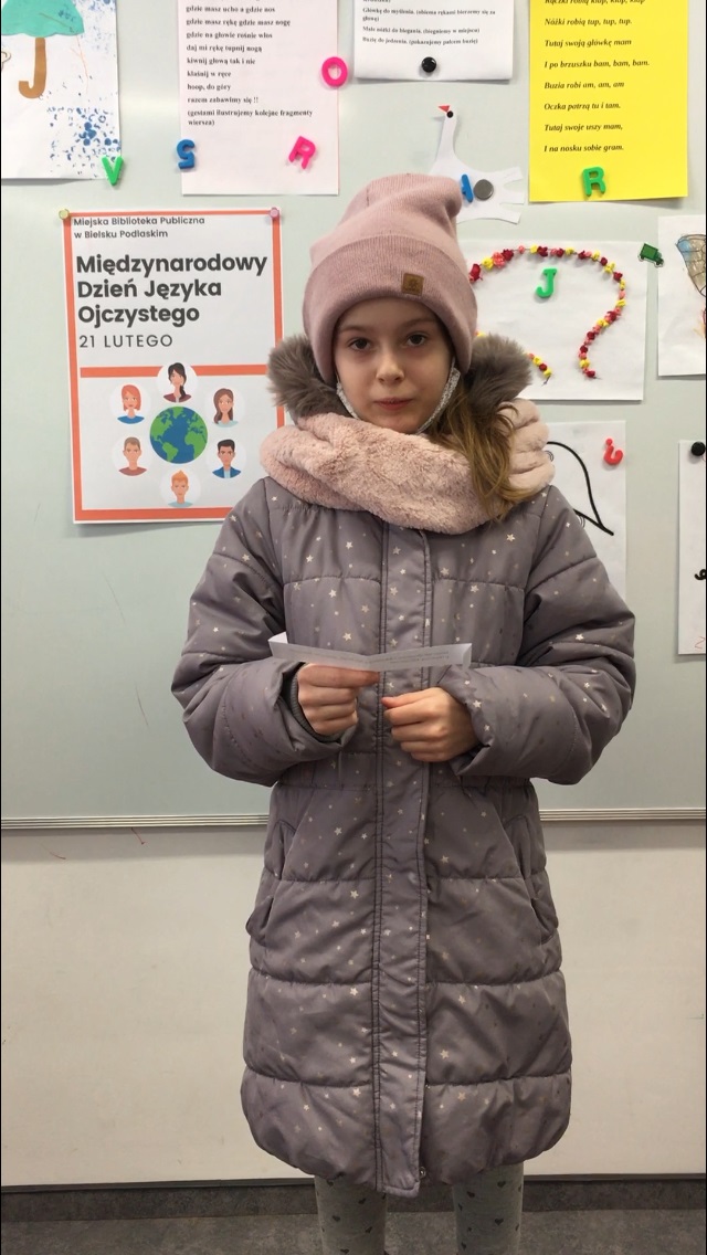 dziewczynka w różowej czapce trzyma kartkę z czytanym wierszem, w tle biała tablica z dziecię-cymi rysunkami i plakatem z napisem: Międzynarodowy Dzień Języka Ojczystego.