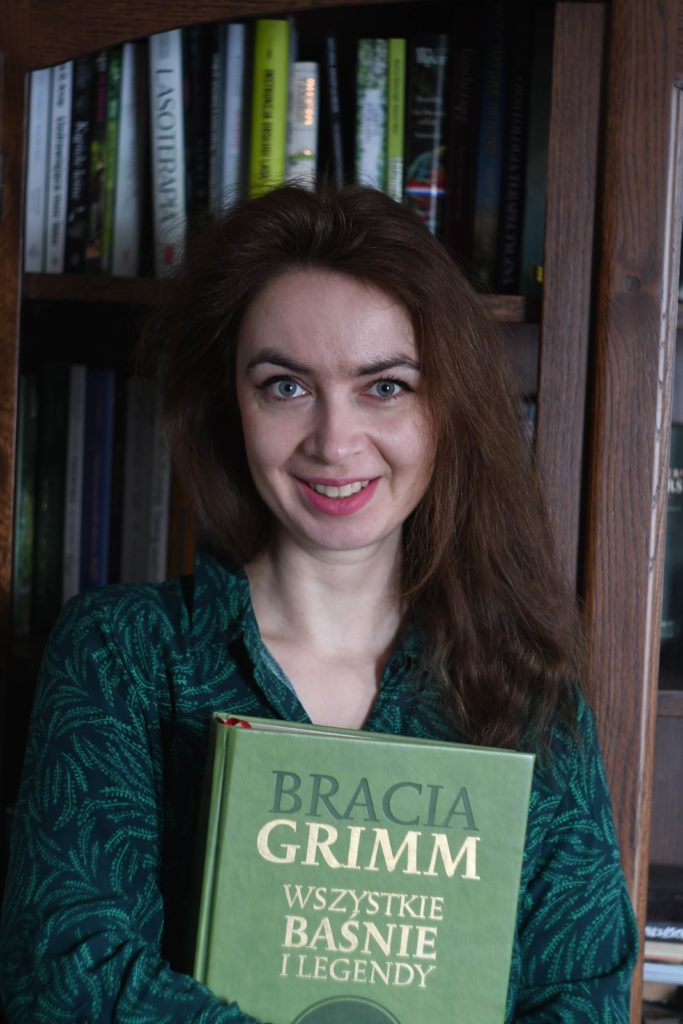 uśmiechnięta kobieta patrzy na wprost trzyma w ramionach książkę widać tytuł Bracia Grimm Wszysktie baśnie i legendy