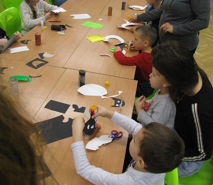 Dzieci siedzą przy stole i wykonują prace plastyczne przy pomocy rodziców