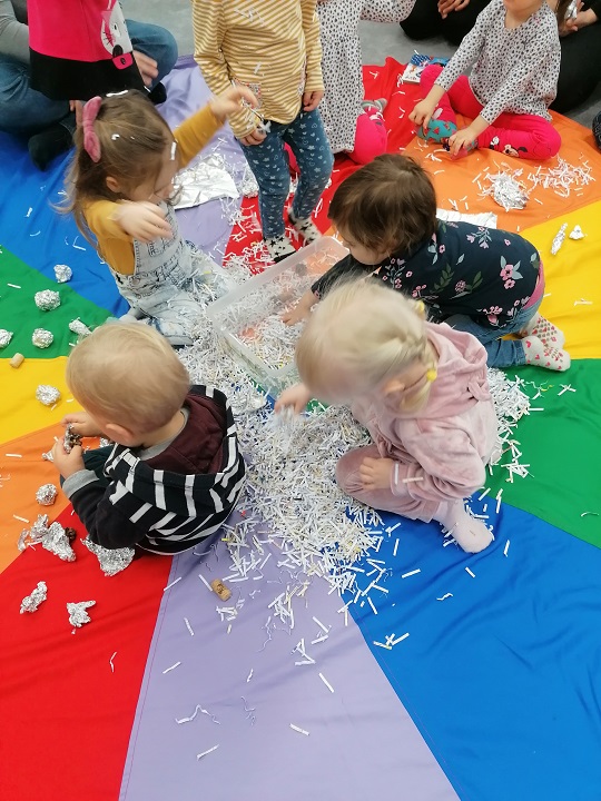 Czwórka dzieci siedzi na podłodze wokół rozsypanych ścinek papieru. Podłoga przykryta chustą w kolorach tęczy.