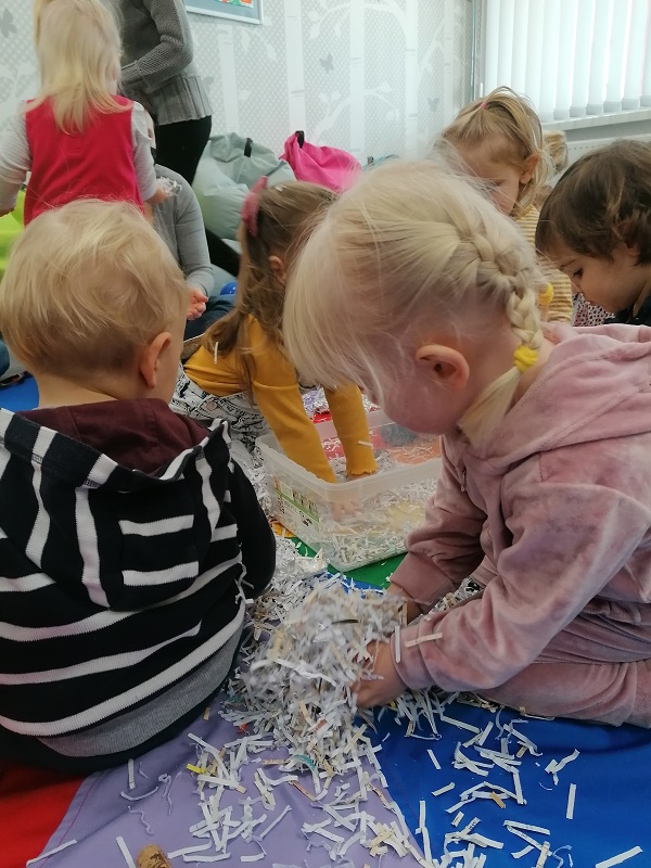 Piątka dzieci siedzi na kolorowej chuście, bawią się ścinkami papieru rozsypanymi na podłodze. Jedna z nich ma ręce włożone do plastikowego pudełka.