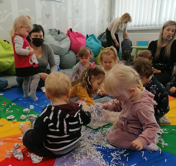 Sześcioro dzieci siedzi na kolorowej podłodze. Pośrodku stoi plastikowe pudełko. Na trzecim planie trzy osoby dorosłe. Dwie siedzą po prawej i lewej stronie. Jedna schylona nad stojącym dzieckiem.