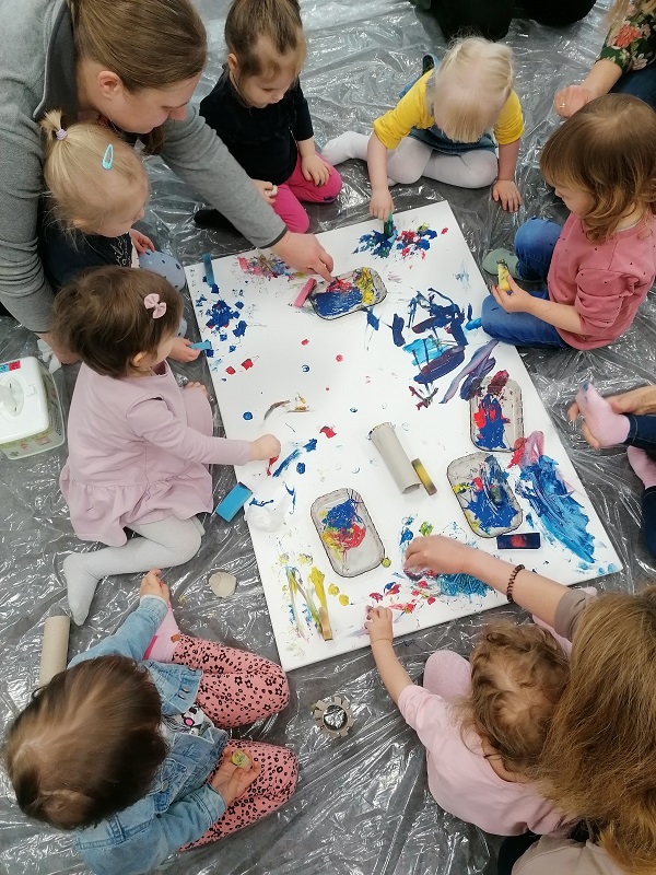 Siedmioro małych dzieci i dorosła osoba z prawej strony siedzą w kole na podłodze, pośrodku między nimi płótno, na którym dzieci malują farbami za pomocą gąbek.