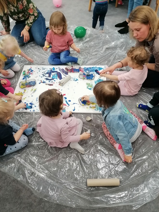 Szóstka dzieci siedzi na podłodze wokół rozłożonego płótna, które malują farbami