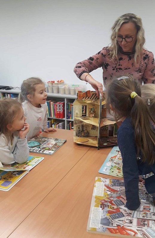 cztery dziewczynki siedzące przy stole nad książkami patrzą na książkę przestrzenną - wiktoriański dom lalek prezentowaną przez panią bibliotekarkę