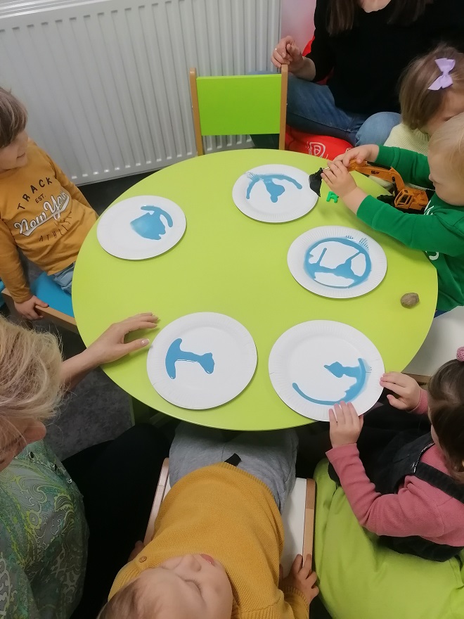 Grupa dzieci siedzi przy stoliku, na którym leży pięć papierowych talerzy z niebieskimi kleksami