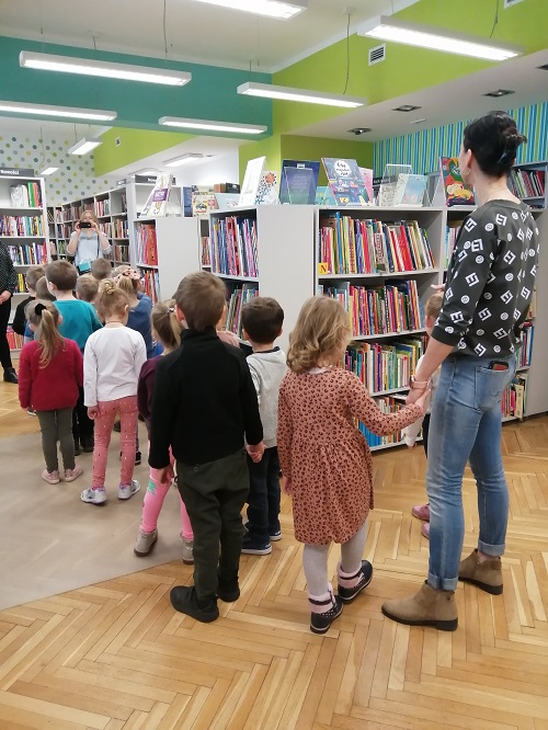 grupa czterolatków wraz z paniami z przedszkola stoi wśród bibliotecznych półek
