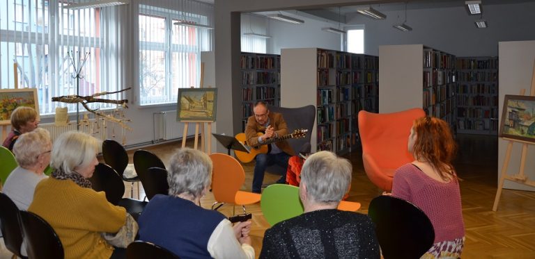 Uczestnicy spotkania siedzą na krzesłach przed nimi mężczyzna gra na gitarze siedząc na fotelu. W tle regały z książkami.