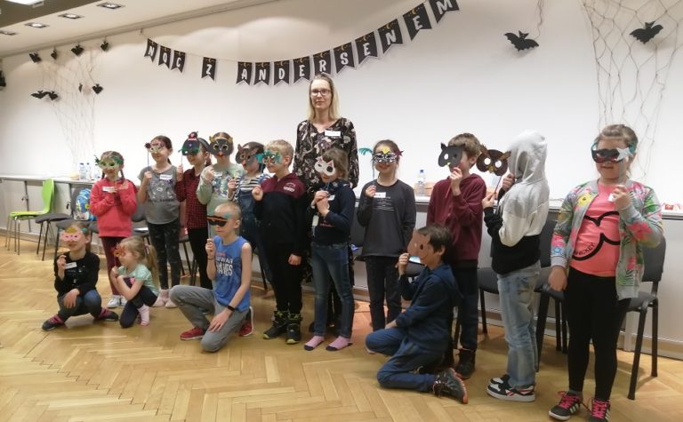 Piętnaścioro dzieci trzymających w dłoniach maski na oczy, ustawionych w dwóch rzędach na tle ściany z napisem Noc z Andersenem. Pośrodku dorosła kobieta - dyrektorka biblioteki