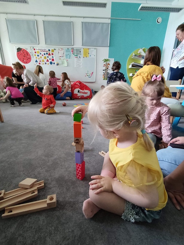 Na piewrwszym planie siedzącą dziewczynka w żółtej bluzce buduje wieże z klocków w tle inne bawiące się dzieci i ich opiekunowie.