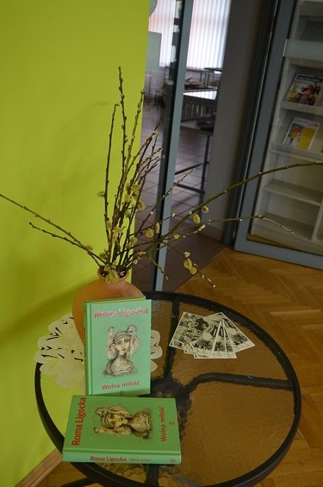 Na szklanym okrągłym stoliku znajduje się wazon z kwiatami i dwa egzemplarze książki Romy Ligockiej „Wolna miłość”.