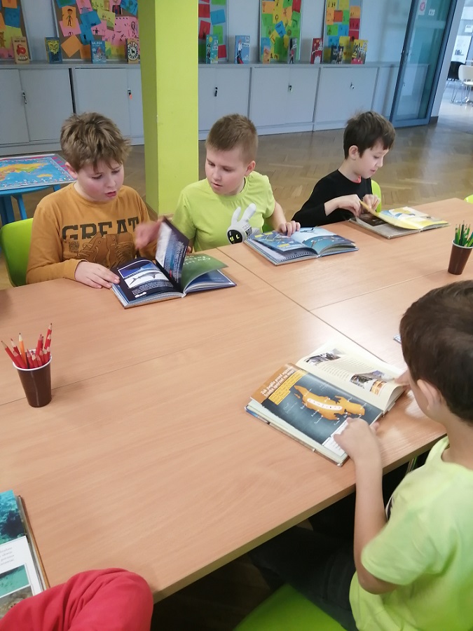 Czterech uczniów siedzi przy stolikach i przegląda książki Neli małej reporterki