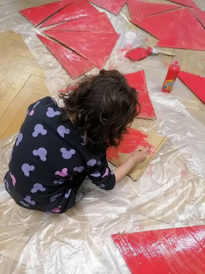 . Dziewczynka z pędzlem w ręku maluje jeden z tekturowych trójkątów. Kolejne leżą wokół niej na podłodze.