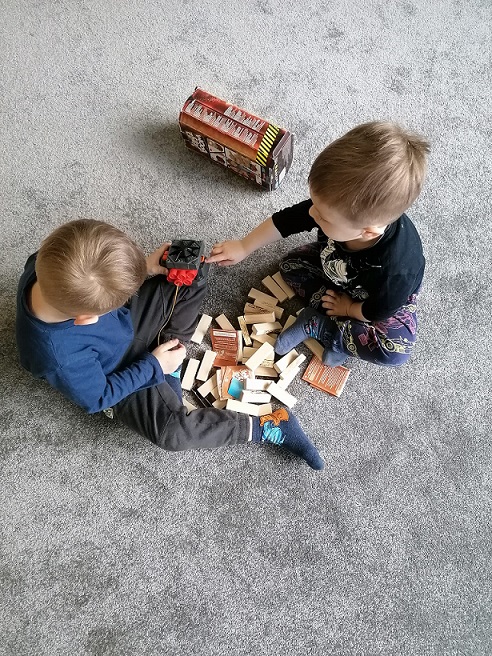 Dwóch chłopców siedzi naprzeciwko siebie na szarym dywanie przed nimi rozsypane klocki.
