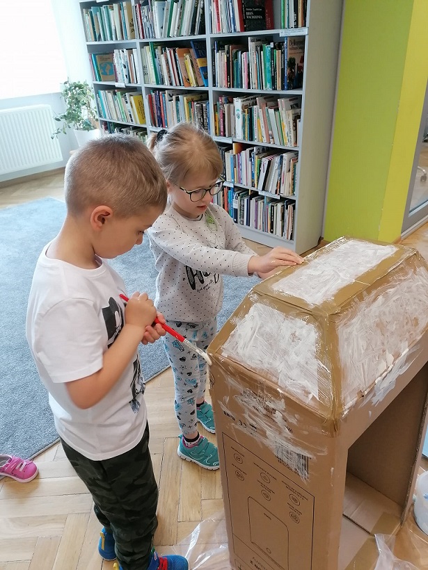Chłopiec z dziewczynka malują farbami kartonowe pudelku za nimi pólka z książkami.