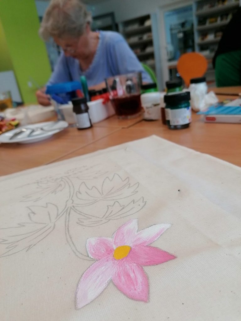 . Na stole torba płócienna z malowanym ręcznie kwiatem. W tle stół z materiałami warsztatowymi przy którym kobieta maluje torbę.