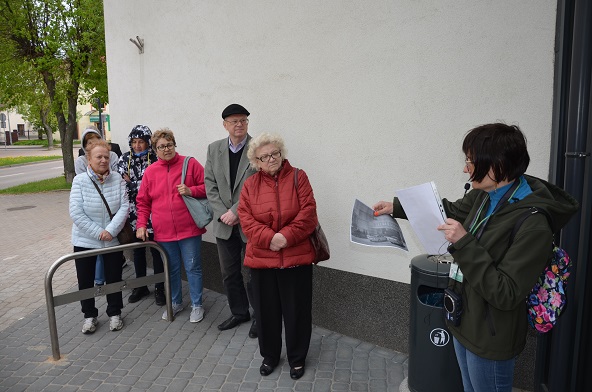 Grupa osób stojących przed BDK od strony kina słucha przewodniczki, która prezentuje stare zdjęcia tego miejsca