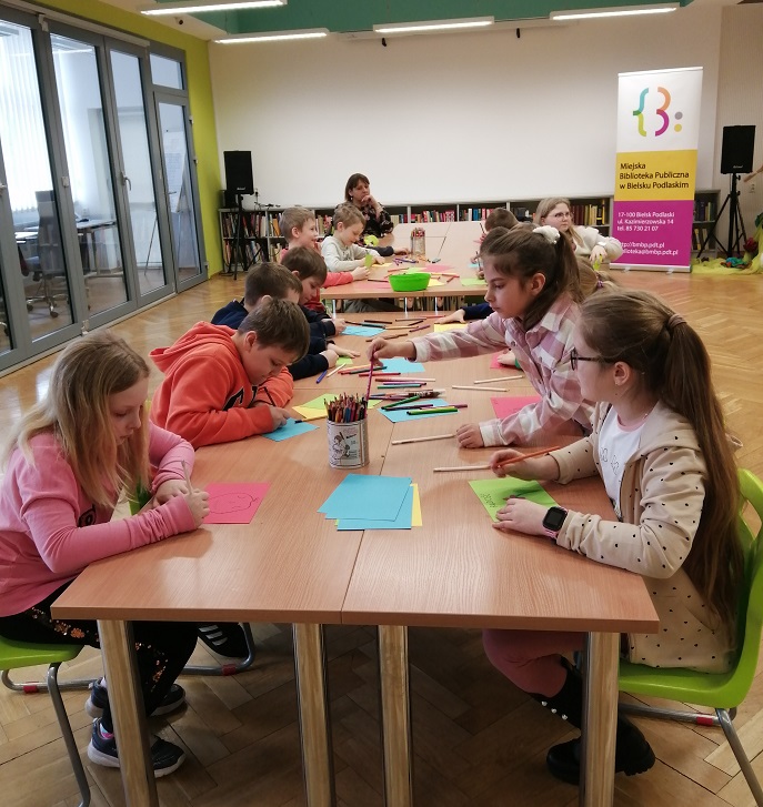 Grupa dzieci siedzi po obu stronach stołu i rysuje po kolorowych kartkach. Na stole leżą kartki, mazaki. W tle z prawej strony widoczny baner biblioteki.