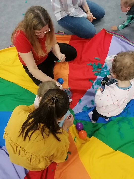 Dwie dorosłe kobiety i dwoje maluchów bawi się na kolorowej chuście Klanza.