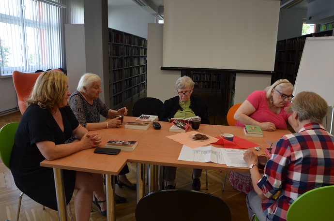 Kilka kobiet siedzi za stołem i dyskutuje przeglądając książki. W tle regały biblioteczne.