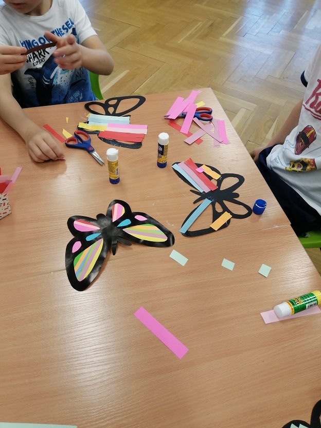 Dzieci pracują nad witrażowymi motylami z papieru, na stole widać częściowo wykonane prace i dziecięce dłonie