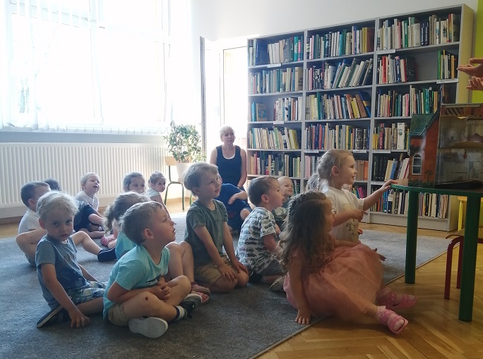 Grupa dzieci siedzi na dywanie (w tle widać panią nauczycielkę) i słucha pani bibliotekarki pokazującej różne rodzaje książek