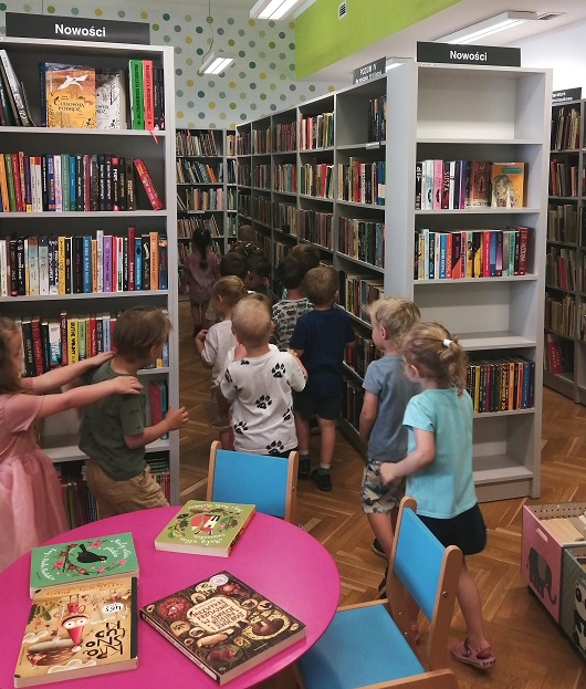 Grupa dzieci tworząc pociąg przechadza się między bibliotecznymi półkami, zwiedza Wypożyczalnię dla Dzieci
