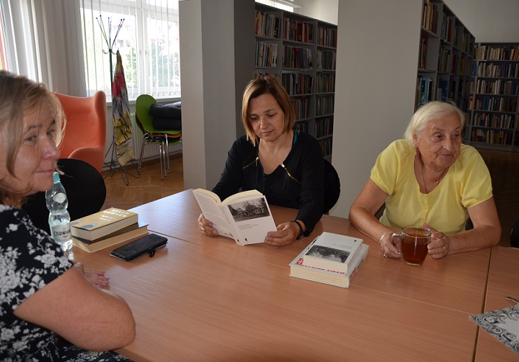 Trzy kobiety siedzą za stołem i dyskutuje przeglądając książki. W tle regały biblioteczne