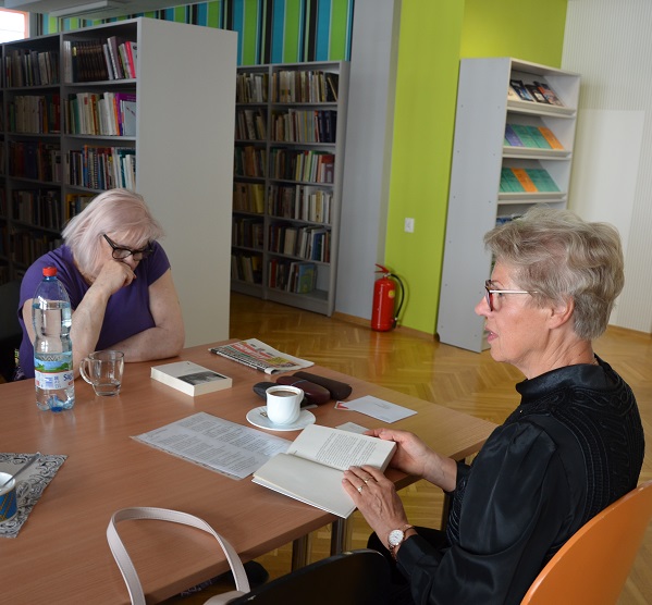 Dwie kobiety siedzą za stołem i dyskutują przeglądając książki. W tle regały biblioteczne