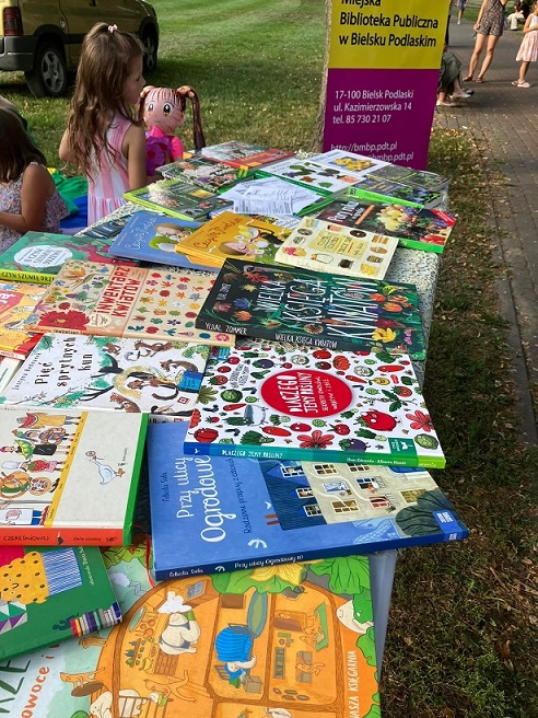 Fragment stołu z wyłożonymi książkami ustawiony w parku w tle fragment chodnika, baneru, auto, a przed nim dziewczynka.