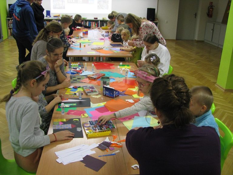 Uczestnicy warsztatów w procesie tworzenia prac plastycznych siedzą wokół długiego stołu, na którym leżą kolorowe kartki, papiery. W tle fragment ściany, a na niej rzucony obraz z ekranu rzutnika