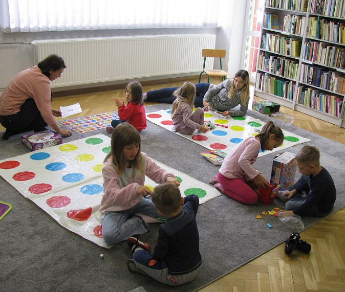 Sześcioro dzieci i dwójka dorosłych grają w planszówki na dywanie w tle regał z książkami.