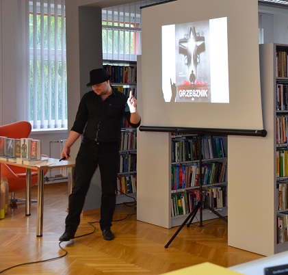 Autor przedstawia prezentację multimedialną żywo gestykulując. W tle stolik, na którym leżą jego książki