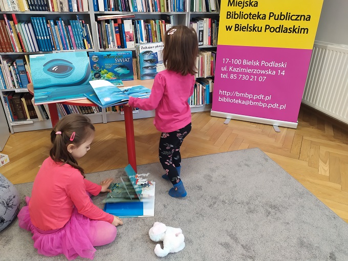 Dwie dziewczynki przeglądają książki o wielorybach. W tle baner z nazwą biblioteki i regał z książkami.