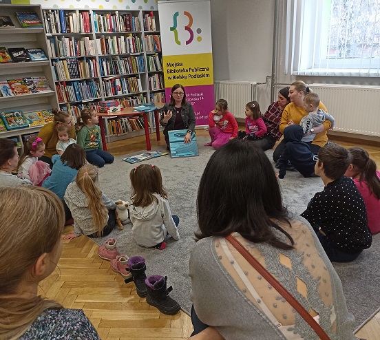 Grupa dzieci i dorosłych siedząca w kręgu na dywanie. W tle baner z nazwą biblioteki i regał z książkami.