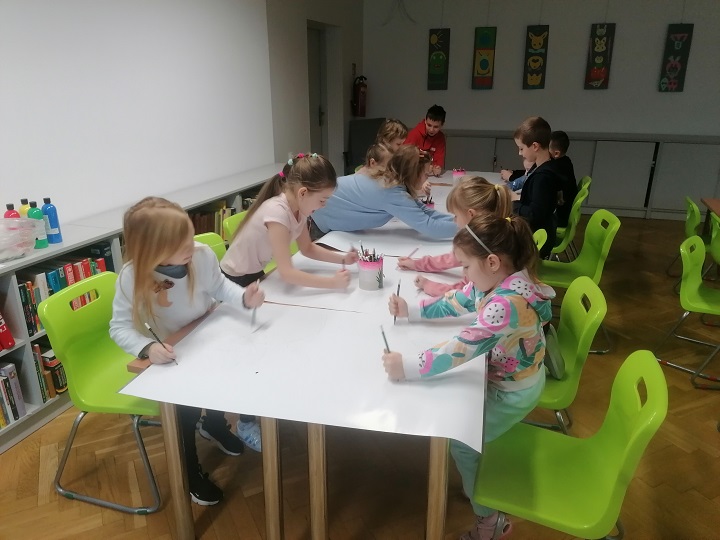 Grupa dzieci przy obu stronach stołu rysuje po nim kredkami.