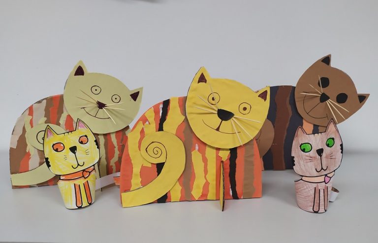 Na półce pięć prac plastycznych, trzy większe papierowe koty w paski i dwa mniejsze.