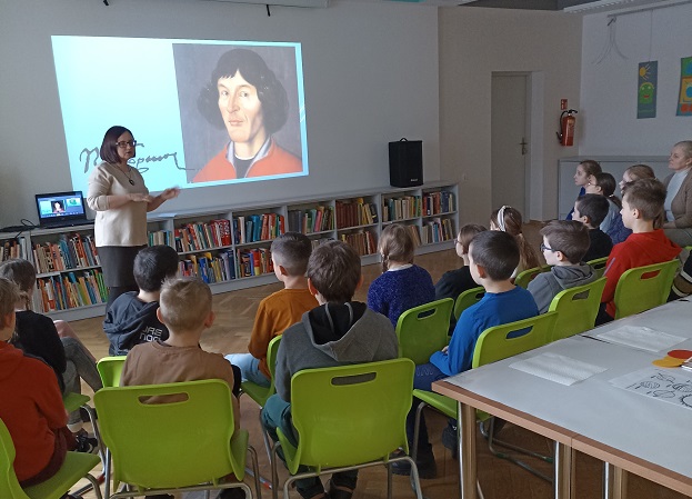 Grupa dzieci siedząca na krzesłach przed ekranem. Na ekranie portret Mikołaja Kopernika