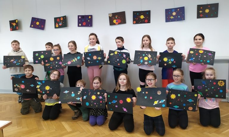 Siedemnaścioro dzieci w dwóch rzędach prezentuje swoje prace plastyczne