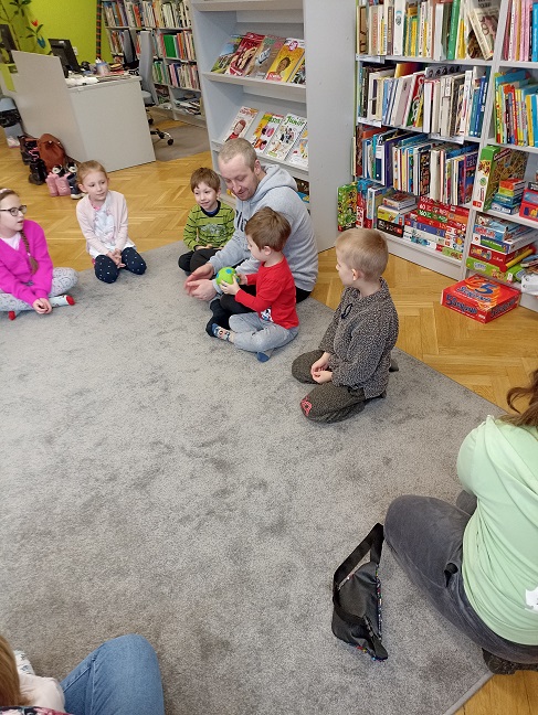 Grupa dzieci siedzi w półokręgu na podłodze, pośród nich mężczyzna w szarej bluzie. Obok niego chłopiec z zieloną piłką w dłoniach. Za nimi regał z książkami
