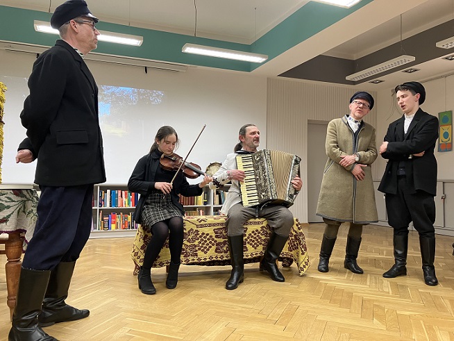 Muzykująca grupa osób w białoruskich strojach ludowych. Trzech mężczyzn śpiewa stojąc. W środku między nimi na przykrytej kapą ławie siedzi grająca na skrzypcach dziewczyna i mężczyzna- akordeonista