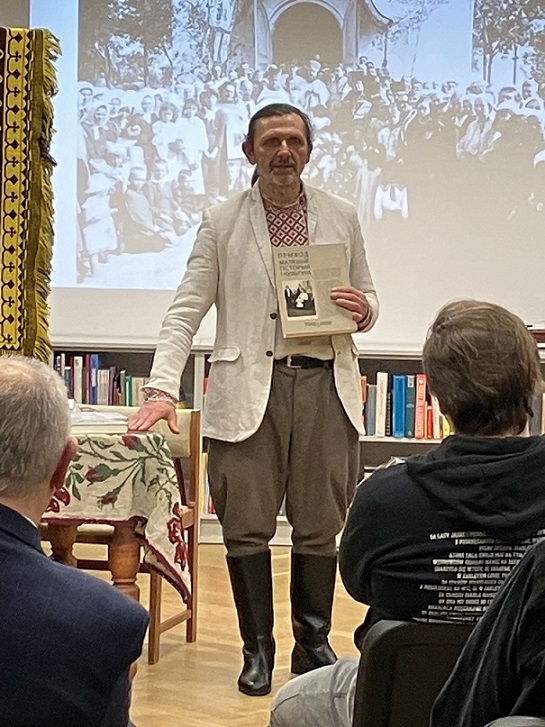 Mężczyzna w białoruskim stroju ludowym prezentuje trzymaną w ręku książkę stojąc na tle ekranu, na którym wyświetlone jest czarno-białe zdjęcie grupy ludzi.