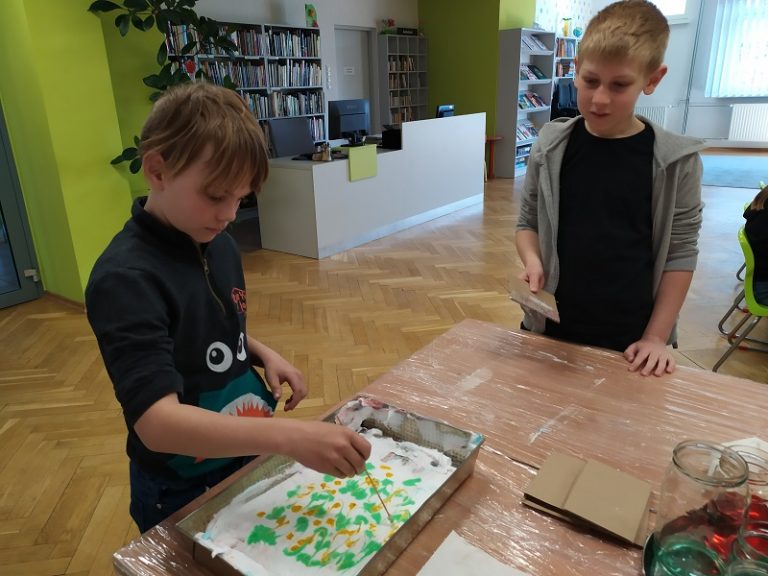 Dwóch chłopców przy stole wykonuje prace plastyczne. Chłopiec po lewej rysuje patyczkiem wzory farbą na masie plastycznej. W głębi regały z książkami i stanowisko bibliotekarza