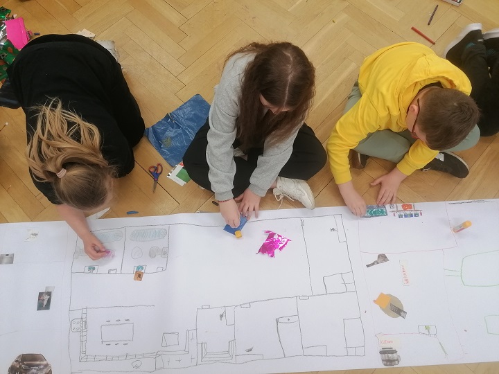 Trzy osoby sfotografowane z góry, siedzą na podłodze przed nimi pas papieru, na którym widoczny jest rysunek z projektem mieszkania