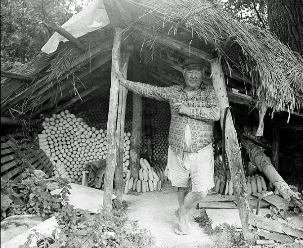 Stojący mężczyzna pod drewnianą wiatą, jedną ręką podpiera słup, patrzy na wprost, z tyłu za nim poukładane w szeregi gomółki mielnickiej gliny.