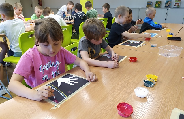 Dziewczynka i czterech chłopców przy długim stole podczas wykonywania pracy plastycznej. Przy użyciu patyczków higienicznych dzieci malują wzory farbą na kartkach. Na drugim planie ośmiu chłopców przy stole w trakcie pracy