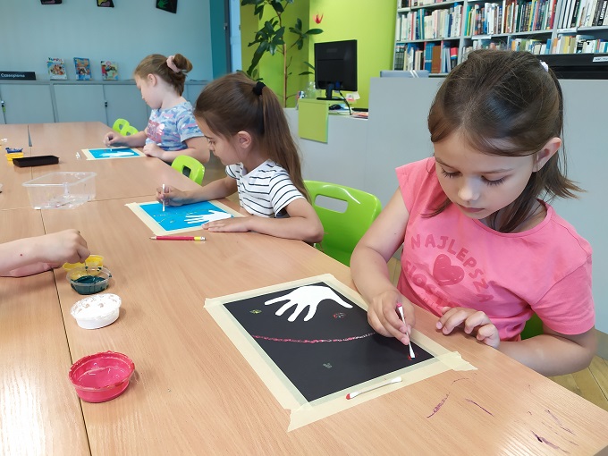 Trzy dziewczynki przy długim stole podczas wykonywania pracy plastycznej. Przy użyciu patyczków higienicznych dzieci malują wzory farbą na kartkach. Na drugim planie stanowisko bibliotekarza i regały z książkami.