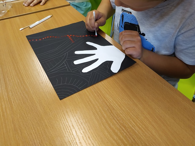 Chłopiec pochylony nad stołem wykonuje pracę plastyczną. Przy użyciu patyczków higienicznych maluje wzory farbą na kartkach.