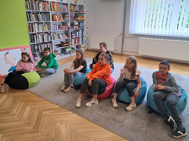Grupa dzieci na kolorowych pufach siedzi w bibliotecznej przestrzeni