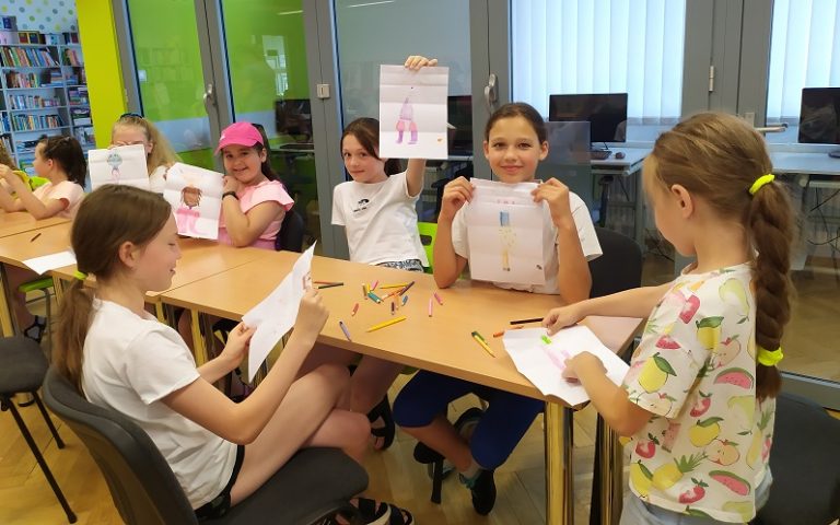 Siedzące przy stole dziewczynki pokazują swoje rysunki. Za nimi przeszklone pomieszczenie z czterema stanowiskami komputerowymi